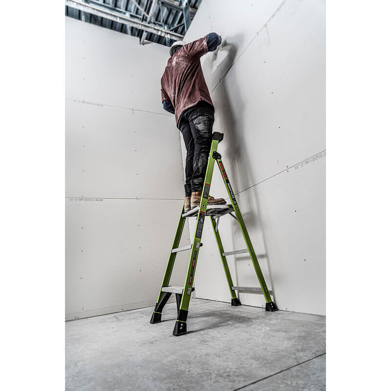 A person stands near the top of a LITTLE GIANT 15395-001 MightyLite IA 5 ft. 300 lb. Rated Fiberglass Stepladder, installing or finishing drywall in a large, unfinished room with high ceilings.