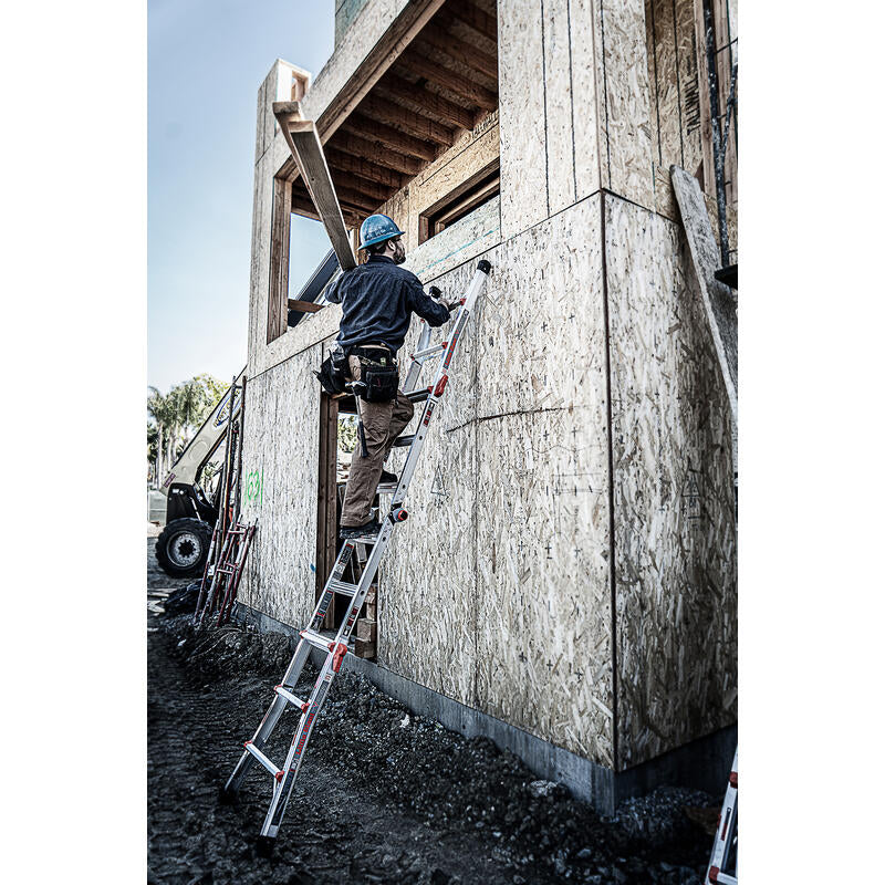 Wearing safety gear, a construction worker climbs a LITTLE GIANT 15413-001 Velocity IA 3 ft. - 11 ft. Aluminum Articulated Extension Ladder leaning against a plywood-framed building at a dirt-covered worksite.
