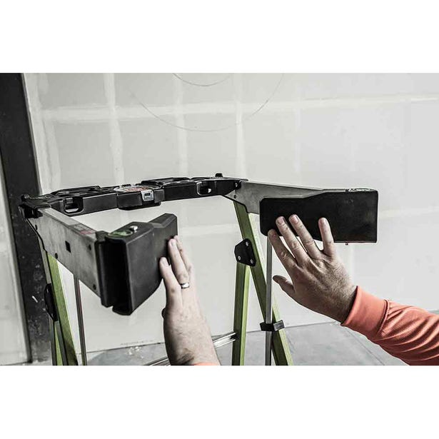 A person adjusts a black accessory atop the LITTLE GIANT 19304 FORTRESS IAA 4 Fiberglass Platform Ladder with Wraparound, set against an unfinished wall—ideal for contractors at any jobsite.