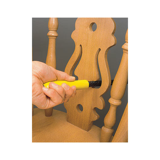 A hand uses the STANLEY 10 IN SURFORM® ROUND FILE with a yellow handle to clean dust from the intricate cut-out design of a wooden chair backrest, highlighting fine woodworking.