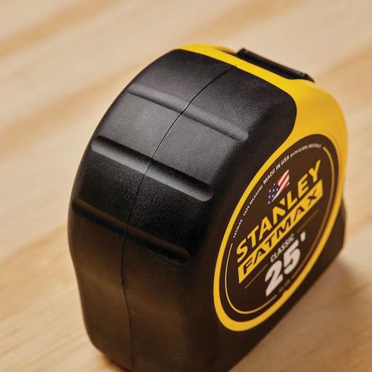 A close-up of a 25 ft STANLEY® FATMAX® Classic Tape Measure, a durable measuring tool with black and yellow casing by STANLEY, resting on a light wooden surface.