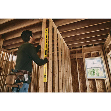 A construction worker uses the Go Build STANLEY® ABS I-Beam Level 1200mm to check vertical alignment of wooden wall framing inside a partially built house, standing in a room with exposed beams and wearing a tool belt.