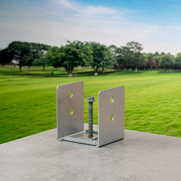 The Simpson Strong-Tie ABU ZMAX® 4-in x 4-in post base bracket, ideal for connecting wood posts to concrete in high-capacity uses, features four holes and a central bolt. It is shown on concrete near a grassy field with trees under a blue sky.