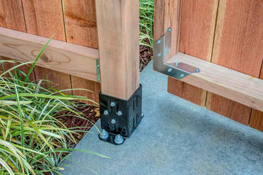 A wooden fence post is anchored to concrete with a Simpson Strong-Tie E-Z Base Black Powder-Coated Post Base for 4x4 lumber and bolts, supporting a wooden gate with metal braces, surrounded by green plants.
