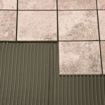 A view of beige square tiles being installed on a surface shows one missing tile, revealing the dark MAPEI Keraflor Thin-Set Tile Mortar in gray with its distinctive ridged texture underneath. The tiles feature a slight marbled pattern and are arranged in a grid, ideal for both interior and exterior floors.