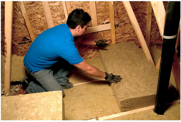 A person in gloves and a blue shirt installs Rockwool RXCB301525 R-30 Comfortbatt 7-1/4 in. x 15 in. x 47 in. fire resistant stone wool insulation batt (29.9 sqft) between attic studs.