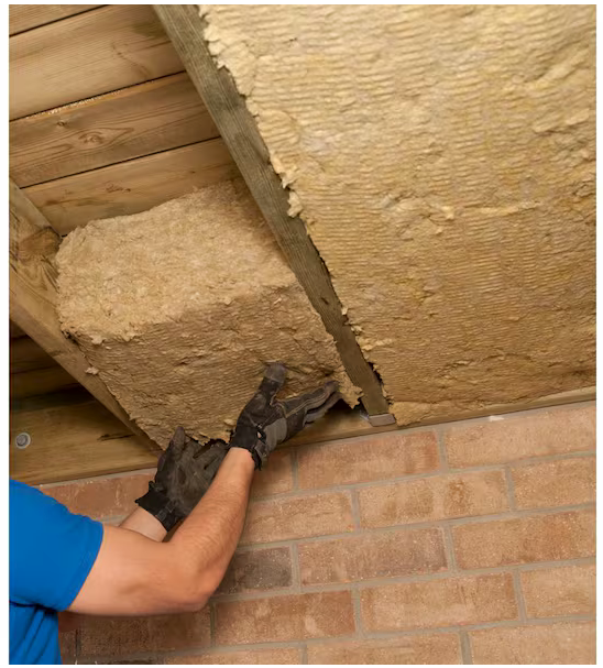 A person wearing gloves installs Rockwool RXCB301525 R-30 Comfortbatt 7-1/4 in. x 15 in. x 47 in. Fire Resistant Stone Wool Insulation Batts between wooden ceiling beams above a brick wall.