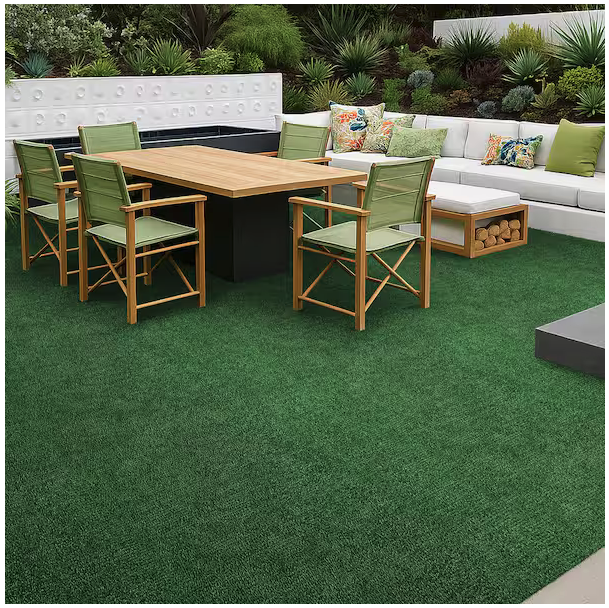 Modern outdoor patio with a pet-friendly TrafficMaster Emerald Green Precut Turf 6 ft. x 8 ft. Artificial Grass Rug, wooden dining table, green cushioned chairs, white sectional sofa with colorful pillows, and lush landscaping with green plants and shrubs.