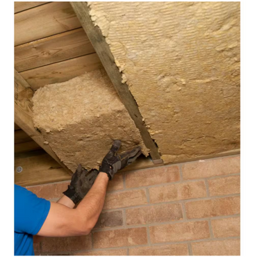 A person wearing gloves installs Rockwool RXCB551525 R23 ComfortBatt 5-1/2 in. x 15.25 in. x 47 in. stone wool insulation panels between wooden ceiling beams above a brick wall.