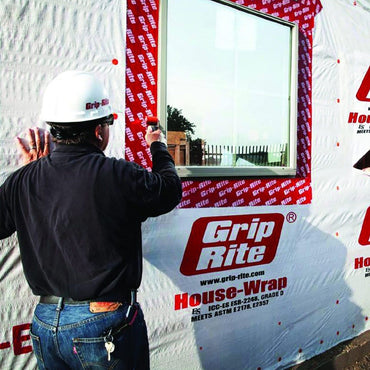 Two workers install a window frame on a building covered in Grip-Rite House-Wrap, sealing it with GRIP-RITE GRWRWDT4 4 in. x 75 ft. self-adhesive elastomer membrane. One wears a white hard hat, and the other has a gray cap, working outdoors in daylight.