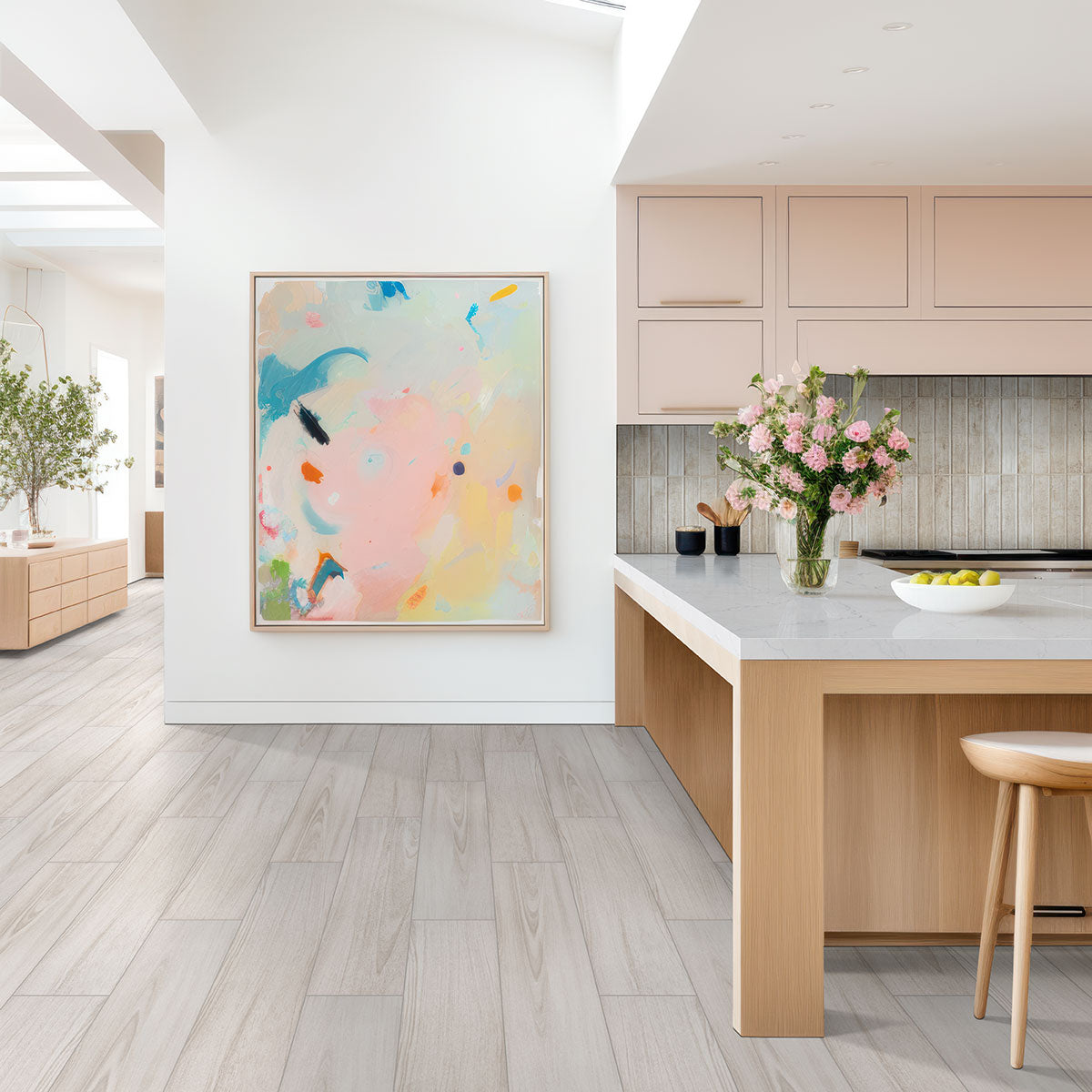 A modern kitchen showcases MSIs Braxton Blanco Porcelain Floor & Wall Tiles, light cabinets, a marble island with pink flowers and a stool, paired with abstract artwork. Large windows allow natural light to fill the space.
