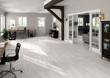 An open-plan room showcases MSIs Braxton Blanco Porcelain Tiles (10 in. x 40 in.) with elegant light wood aesthetics and modern decor. A desk, chair, large windows, and glass doors define the space, allowing natural light to highlight its minimalist design with expansive tile planks providing an elegant touch.