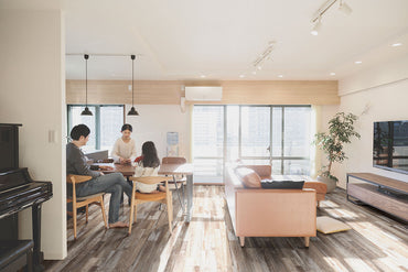 A family of three sits at a dining table in a bright living room with large windows, cozy furnishings, light wood tones, a piano, and warm sunlight. The space features MSI Cyrus Bembridge Luxury Vinyl Planks (23.77 sq ft) waterproof flooring.