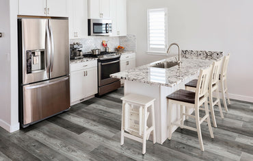 Modern kitchen with stainless steel appliances, white cabinets, granite counters, and a large island with sink. Three white barstools line the island, while MSI Cyrus Boswell Luxury Vinyl Planks bring style and charm to the floor.