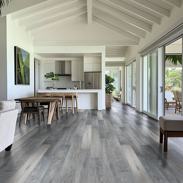 A modern, open-plan living and dining area showcases MSI Cyrus Finely Luxury Vinyl Planks (23.77 SQFT/box), featuring gray and greige wood tones, white walls, vaulted ceiling with beams, a minimalist kitchen, and glass doors to a lush patio.