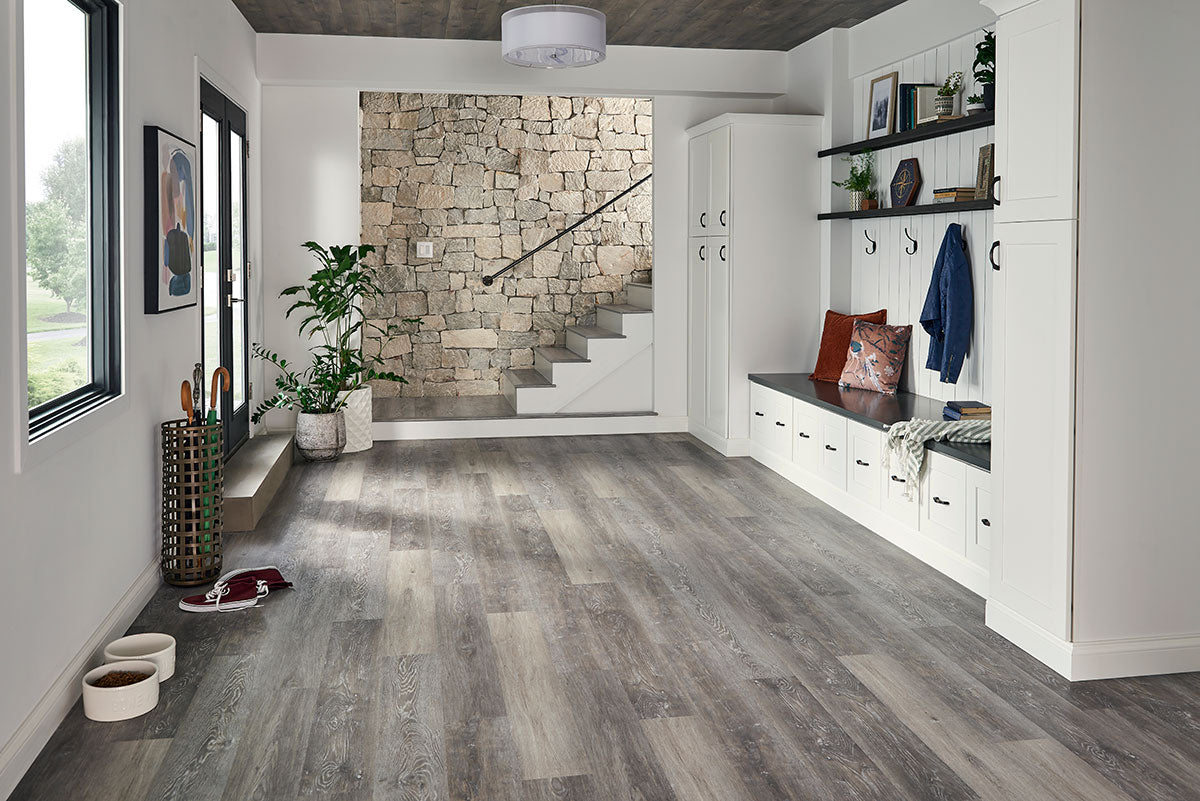 A modern mudroom features MSI Cyrus Finely Luxury Vinyl Planks BOX (23.77 SQFT) in gray and greige wood tones, with white built-in cabinets, benches, storage hooks, shelves, a stone accent wall, staircase, shoes, and pet bowls on the waterproof flooring.