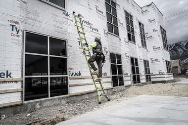 A construction worker in safety gear climbs a LITTLE GIANT 17117 CONQUEST 2.0 1A 7.5-15 F/G ARTICULATED EXTENSION LADDER against a building with Tyvek-wrapped exterior walls under construction.