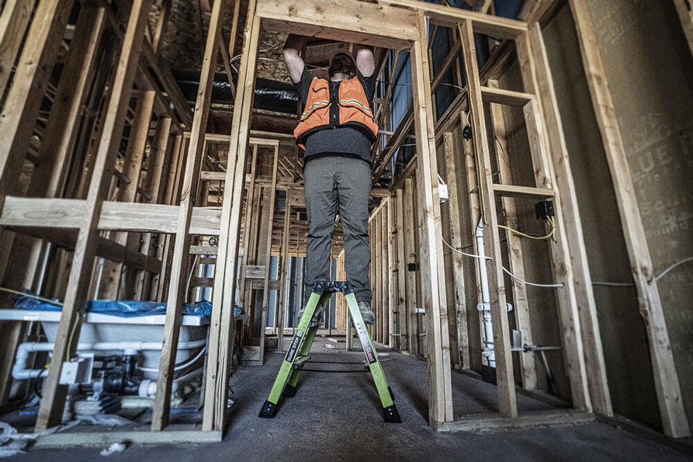 A construction worker in an orange safety vest stands on a LITTLE GIANT 11953 Sure Step 1AA 3-Step Double-Sided Fiberglass Step Stool, working on the framing of a building with exposed wooden beams and unfinished walls.