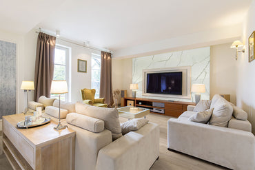 A spacious, modern living room with beige sofas, a glass coffee table, a green armchair, floor lamps, MSI Cyrus Sandino Luxury Vinyl Planks flooring (waterproof SPC core), a wall-mounted TV, and windows with curtains letting in natural light.