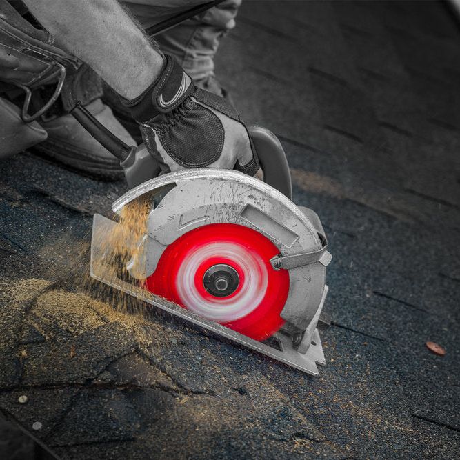 A worker expertly uses the DIABLO D0724DA 7-1/4 in. x 24-Teeth Demo Demon™ saw blade by Diablo with a distinct red edge to cut through a black shingle roof, demonstrating its precision. Sawdust scatters as the worker, in protective gloves, skillfully handles the task against a mostly monochrome backdrop.
