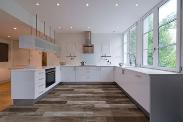 Modern kitchen with white cabinets, stainless steel appliances, and large windows features MSI Cyrus Weathered Brina Luxury Vinyl Planks; these 100% waterproof floors provide durable style and a rustic wood look beneath natural light.