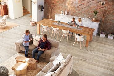 A cozy living and dining room features MSI’s XL Cyrus Akadia Luxury Vinyl Planks (22.44 SQFT) with neutral beige tones, brick walls, and warm decor. Two people chat on a couch while two others work at a long dining table.