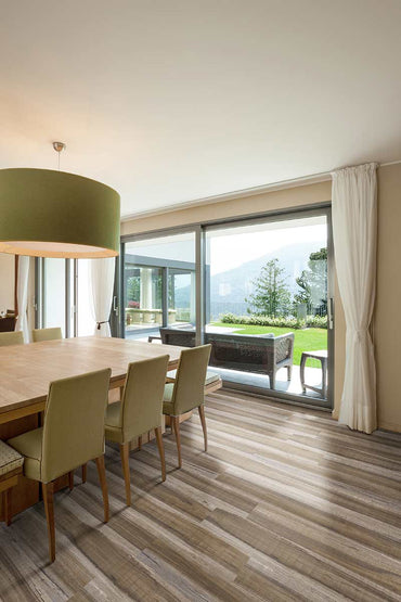 Modern dining room with wood table, green chairs, and CrystaLux™ pendant light. Sliding glass doors reveal patio seating and scenic views, enhanced by MSI XL Cyrus Exotika Luxury Vinyl Planks for a touch of elegance indoors.