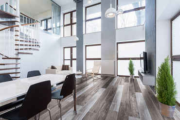 Modern living room with large windows, high ceilings, a wooden spiral staircase, white table with black chairs, sofa, armchair, TV, and two potted plants on MSI XL Cyrus Weathered Brina Luxury Vinyl Planks (22.44 SQFT/box).
