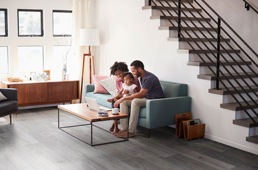 A family of three relaxes on a teal sofa in a stylish living room with MSI XL Cyrus Woburn Abbey Luxury Vinyl Planks flooring, enjoying time together by the coffee table. The space features large windows, a staircase, and mid-century décor.