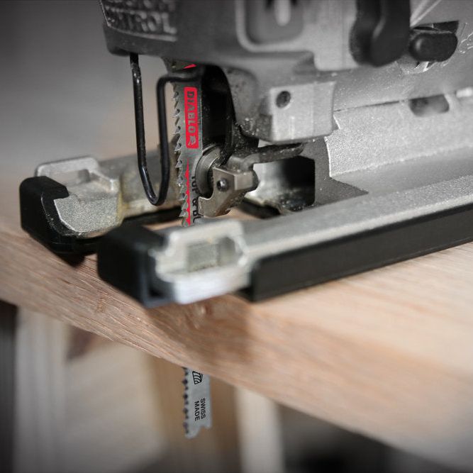 A DIABLO DJT308BFP5 4-5/8 in. 13TPI Bi-Metal Jig Saw Blade effortlessly cuts a wooden plank on a workbench, with its ultra-hardened edge partially inserted. The blurred background highlights the precision of this Diablo tool.