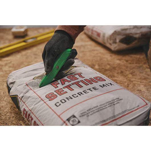 A gloved hand uses a STANLEY® Hi-Visibility Retractable Knife to open a Fast Setting Concrete Mix bag on a work surface, with another bag and a yellow level nearby.