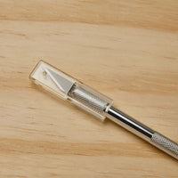 The STANLEY Hobby Knife, featuring a textured metal handle, replaceable blades, and a clear plastic safety cap, rests on a light wooden surface.