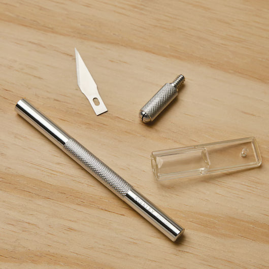 A disassembled STANLEY Hobby Knife, featuring a metal handle, replaceable blades, blade holder, and clear plastic safety cap, is laid out on a light wooden surface.