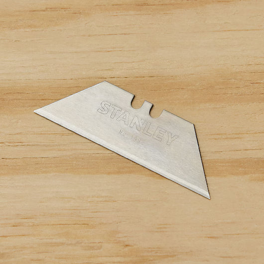 A single STANLEY 1992® Heavy-Duty Utility Blade from a 5 PK lies on a light wooden surface. The rectangular blade features angled edges and a notched top for easy replacement in utility knives.