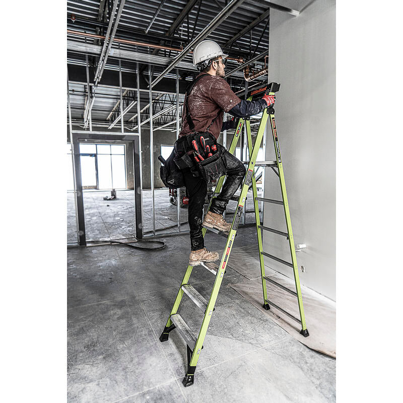 A construction worker in a helmet and tool belt stands on a LITTLE GIANT 15398-001 MightyLite IA 8 ft. 300 lb. fiberglass stepladder inside a building with exposed beams and unfinished walls.
