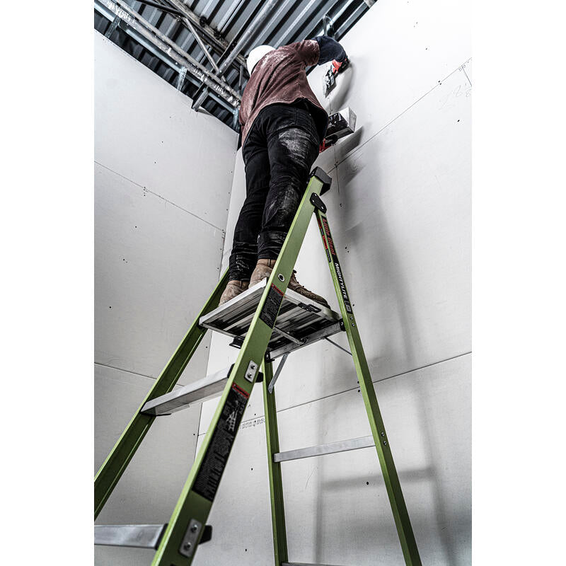 A worker in a hard hat stands on a LITTLE GIANT 15394-001 MightyLite IA 4 ft. 300 lb. fiberglass stepladder, installing or adjusting cables on a high white wall in an unfinished building with exposed pipes and beams.