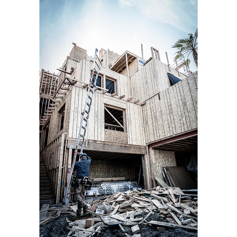 A construction worker stands beside a LITTLE GIANT 15422-001 Velocity IA 7 ft. - 19 ft. Aluminum Articulated Extension Ladder, which is leaning against a partially built wooden house amid scattered wood planks and construction materials.