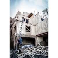 A construction worker stands beside a LITTLE GIANT 15422-001 Velocity IA 7 ft. - 19 ft. Aluminum Articulated Extension Ladder, which is leaning against a partially built wooden house amid scattered wood planks and construction materials.