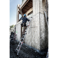 Wearing safety gear, a construction worker climbs a LITTLE GIANT 15413-001 Velocity IA 3 ft. - 11 ft. Aluminum Articulated Extension Ladder leaning against a plywood-framed building at a dirt-covered worksite.