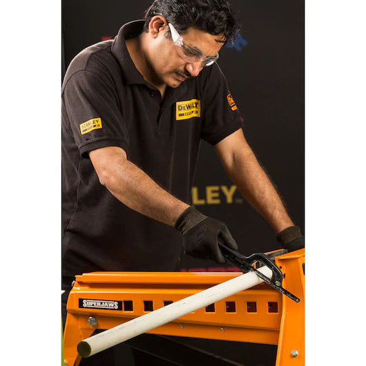 A man in a black DeWalt polo shirt, wearing safety glasses and gloves, uses the STANLEY® Mini Hacksaw to cut a white pipe secured on an orange workbench.