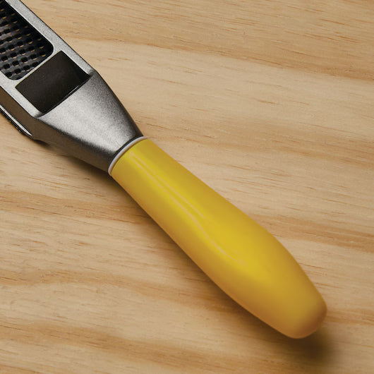 A STANLEY® SURFORM FLAT FILE - REGULAR CUT 10IN (250MM), resembling a metal garlic press with a yellow handle, rests on a light wooden surface.