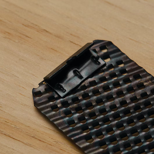 A close-up of a STANLEY 5-1/2 IN SURFORM® POCKET FINE CUT REPLACEMENT BLADE with a grid pattern and dark, iridescent finish rests on a light wooden surface, its partially open end echoing classic woodworking tool design.