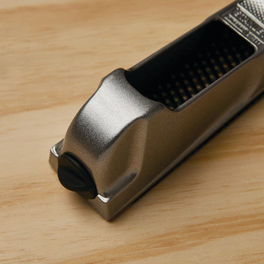 A close-up of the STANLEY 6 IN SURFORM® POCKET PLANE, a versatile metal shaping tool with an attached container, is shown on a light wooden surface.