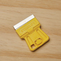 A STANLEY 1-3/16 in High Visibility Mini-Razor Blade Scraper with a yellow handle and metal blade sits on a light wood surface.