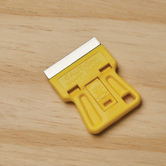 A STANLEY 1-3/16 in High Visibility Mini-Razor Blade Scraper with a yellow handle and visible metal blade rests on a wooden surface, displaying the text “CAUTION: SHARP BLADE.”.
