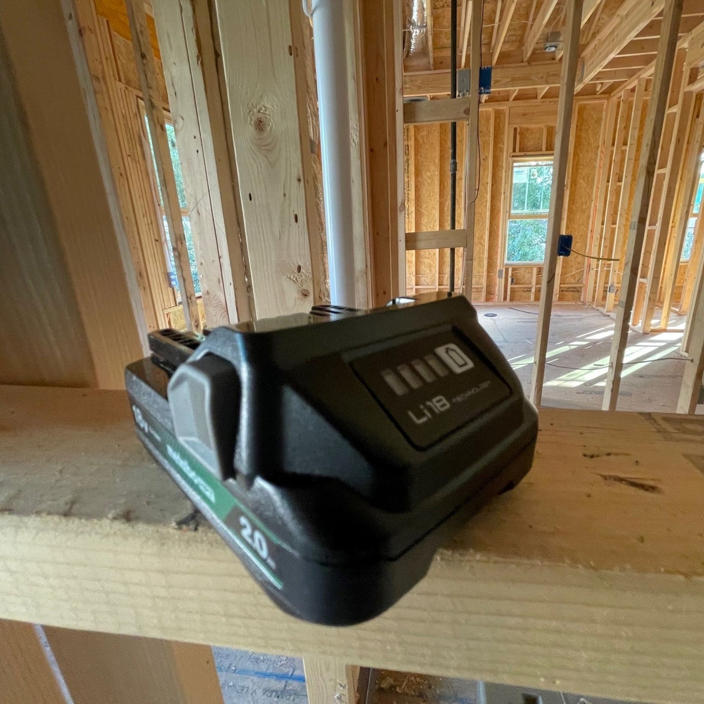 A close-up of a Go Build Metabo HPT 377797M BSL1820M 18V 2.0Ah Lithium Ion Battery with Fuel Gauge on a wood beam inside a sunlit, unfinished house with exposed framing in the background.