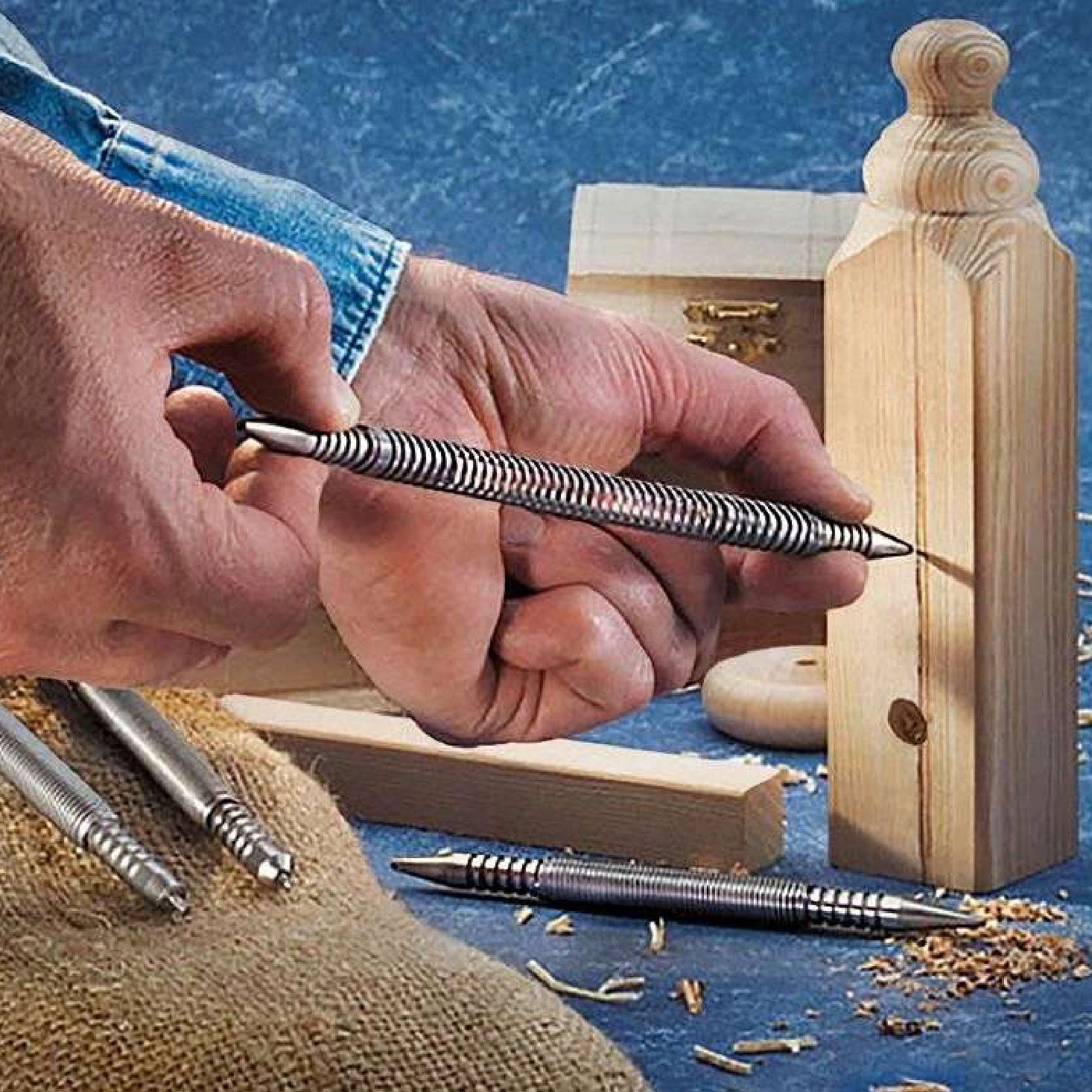 A person uses the SPRING TOOLS 32R12-1 Combination Center Punch and Nail Set to mark a wooden block. Other SPRING TOOLS metal scribe tools, a center punch, wood shavings, and wood pieces are visible on a blue surface in the background.
