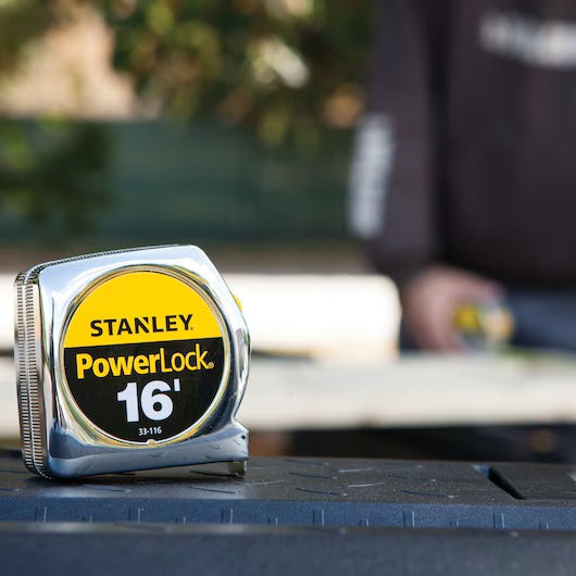 A close-up of a STANLEY 16 ft PowerLock® Tape Measure on a work surface, with a blurred background showing someone in work attire at an outdoor workspace. This reliable tool is trusted for accurate measurements on any project.