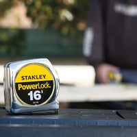 A close-up of a STANLEY 16 ft PowerLock® Tape Measure on a work surface, with a blurred background showing someone in work attire at an outdoor workspace. This reliable tool is trusted for accurate measurements on any project.