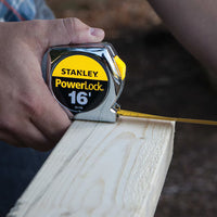 A person uses the STANLEY 16 ft PowerLock® Tape Measure, extending the tape to measure a piece of light-colored wood.
